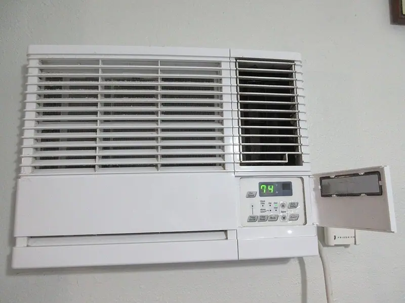 A window type air conditioner fitted into a wall opening, seen from inside the room, with its digital display reading 74 and a hinged panel open at the right-hand end.
