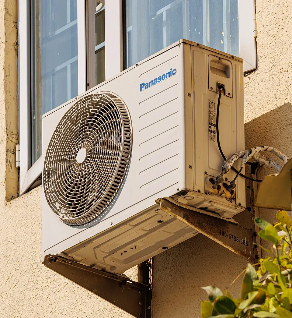 A Panasonic outdoor air conditioning unit on a rusting wall bracket beside a window, its pipework taped and exposed.